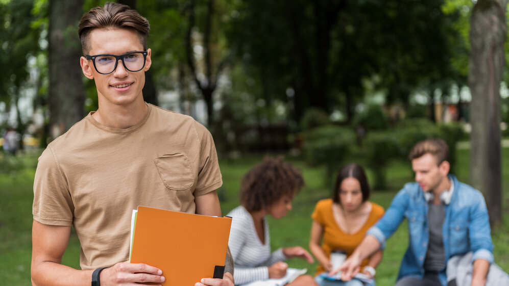 Zweiter Bildungsweg Studium: Das Foto zeigt einen jungen Mann mit Schulunterlagen in der Hand vor einer Gruppe von drei anderen Schülern bzw. Studenten, womit symbolisiert werden soll, dass diese auf dem zweiten Bildungsweg die gymnasiale Matura nachholen, um später an der Universität mit dem Studium zu beginnen.